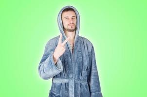 Portrait of young caucasian bearded man in blue bathrobe shows victory or peace gesture isolated on green background photo