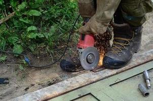 Rectificado eléctrico de ruedas en estructura de acero al aire libre. viejo trabajador manual con guantes protectores cortando puertas de metal con molinillo foto