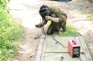 Old man welder in brown uniform, welding mask and welders leathers, weld metal door with arc welding machine outdoors photo