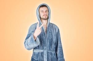 Portrait of young caucasian bearded man in blue bathrobe shows victory or peace gesture isolated on orange background photo