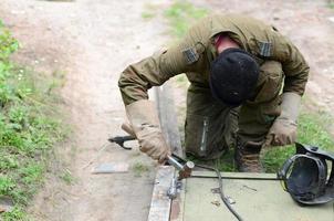 Old man welder in brown uniform prepares metal door surface for welding with arc welding machine outdoors photo