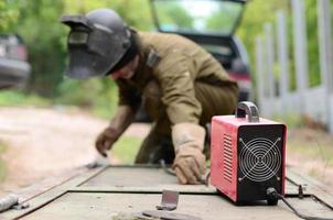 Old man welder in brown uniform, welding mask and welders leathers, weld metal door with arc welding machine outdoors photo