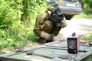 Old man welder in brown uniform, welding mask and welders leathers, weld metal door with arc welding machine outdoors photo