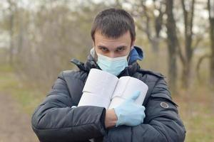 Covidiot concept. Young man in protective mask holds many rolls of toilet paper outdoors in spring wood photo