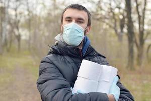 Covidiot concept. Young man in protective mask holds many rolls of toilet paper outdoors in spring wood photo