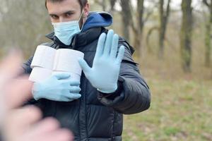 Covidiot concept. Young man in protective mask holds many rolls of toilet paper and shows stop gesture outdoors in spring wood photo