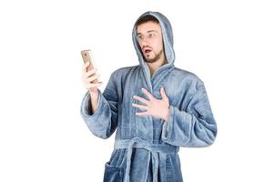 Portrait of young caucasian bearded man in blue bathrobe shows surprised emotion and watch to smartphone screen isolated on white background photo