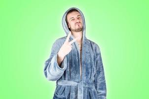 Portrait of young caucasian bearded man in blue bathrobe shows victory or peace gesture isolated on green background photo