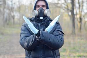 Portrait of young man in protective gas mask and rubber disposable gloves shows stop gesture outdoors in spring wood photo