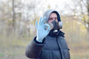 Portrait of young man in protective gas mask and rubber disposable gloves shows okay gesture outdoors in spring wood photo