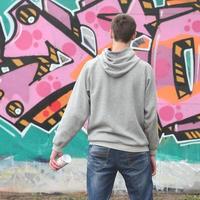 A young graffiti artist in a gray hoodie looks at the wall with photo