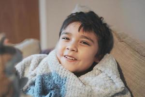 retrato de estilo de vida niño mirando la cámara con un niño sonriente y feliz acostado en un sofá relajándose en la mañana en invierno, niño cómodo acostado en el sofá, concepto de niños positivos foto