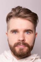 Close-up portrait of young male with beard and mustache and trendy hairdo. Wears casual hoodie, has serious expression in studio against white background photo