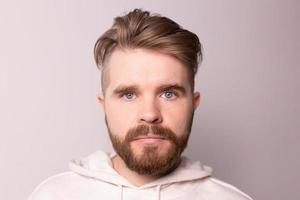 Portrait of young male with beard and mustache and trendy hairdo. Wears casual hoodie, has serious expression in studio against white background photo