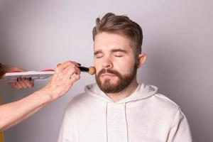 Bearded man getting makeup. Hand of visagist using brush. photo