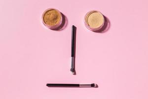 cara divertida con pinceles de maquillaje y polvo mineral sobre fondo rosa con espacio para copiar, vista superior. concepto de belleza y humor cosmético. foto