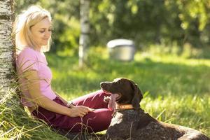 retrato de una joven rubia sentada, relajándose en el césped cerca de un árbol en el parque con un perro de caza de la raza kurz-haar. vacaciones de verano foto