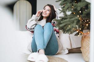 Young beautiful asian woman with dark long hair in cozy sweater using mobile smartphone on bed in room with Christmas tree at home photo