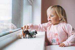 niña pequeña jugando con juguetes de animales en el alféizar de la ventana en la habitación de los niños en casa foto