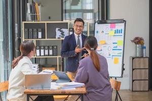 equipo de negocios tres hombres y mujeres asiáticos sonrientes y felices se unen usando tabletas y computadoras portátiles para intercambiar ideas y trazar estadísticas en un tablero de gráficos corporativos dentro de la empresa. concepto de colegas. foto