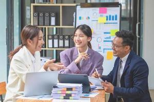 equipo de negocios tres hombres y mujeres asiáticos sonrientes y felices se unen usando tabletas y computadoras portátiles para intercambiar ideas y trazar estadísticas en un tablero de gráficos corporativos dentro de la empresa. concepto de colegas. foto