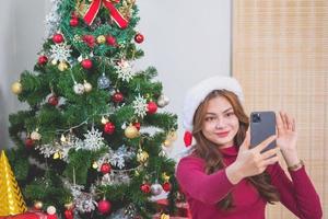 Merry Christmas and Happy Holidays A young woman with a beautiful face in a red shirt shows joy with gift boxes in a house with a Christmas tree decorated with a Christmas tree.  Portrait before Xmas photo