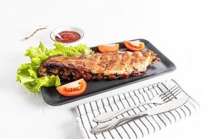 Grilled pork ribs are served on a rectangular dark ceramic plate with fresh vegetables and a bowl of sauce. white background. recipe menu. photo