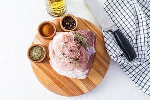 a large fresh piece of pork for baking lies on a round wooden board with various spices. top view. white background. photo