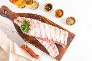 top view of fresh pork ribs lying on a wooden long cutting board with spices in bowls on a white background.