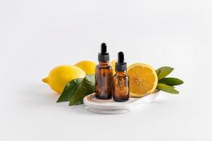 two cosmetic bottles with a pipette with a product based on lemon essential oil stand on a white plaster tray among lemons and leaves. photo