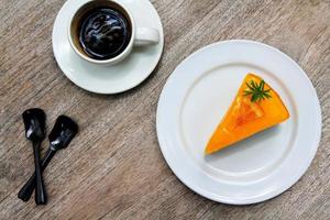 pastel de naranja de primera vista en plato o plato blanco, taza blanca de café negro y cuchara de plástico sobre fondo de madera con espacio para copiar. endecha plana de comida dulce o de postre en la mesa de madera. juego de bocadillos para comer. foto