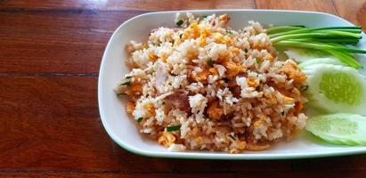 Thai fried rice with pork, fresh vegetable , slices of cucumber and spring onions on white plate or dish on wooden table with copy space on left at Thai food restaurant. Asian food for lunch. photo