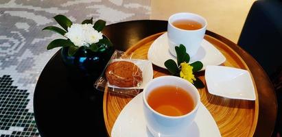 taza blanca de té caliente con coco o galleta de chocolate y flor en una bandeja de madera marrón sobre la mesa. bebida caliente y merienda con concepto de servicio de bebida de bienvenida. foto