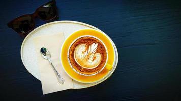 Top view of cup of art cappuccino or latte coffee putting in tray with tea spoon, napkin and brown sunglasses on blue wooden background. Flat lay of hot drinking with copy space on right. photo