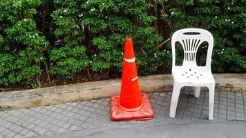 el cono de tráfico naranja y la silla de plástico blanca están en el sendero con flores blancas y plantas verdes o fondo de árbol en el jardín con espacio para copiar. asiento con objeto de canto de tráfico. foto