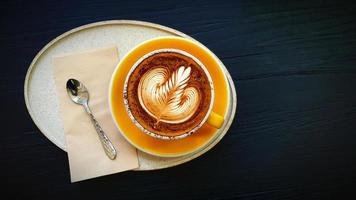 Top view of cup of art cappuccino or latte coffee putting in tray with tea spoon and napkin on dark blue wooden background. Flat lay of hot drinking with copy space on right. Food design concept. photo