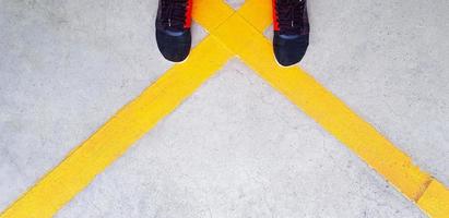 Top view of feet wearing red and back sneakers or fashion shoes on yellow cross line and concrete floor or road with copy space. Flat lay of body part of human stand on street with traffic sign. photo