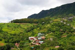 Many house or home built arrange on green mountain with sky background. Village on hill at North of Thailand. Structure of building among tropical forest or jungle. People live in nature. Landscape photo