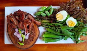 Top view of Thai local food with fried or grilled pork, fermented fish, red onions and green chili on wooden cutting board, boiled egg and fresh vegetable on white plate or dish. Spicy Asian food photo