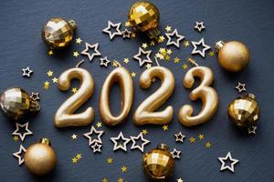 las figuras doradas 2023 hechas de velas sobre un fondo de pizarra de piedra negra están decoradas con una decoración festiva de estrellas, lentejuelas, ramas de abeto, bolas y guirnaldas. tarjeta de felicitación, feliz año nuevo. foto