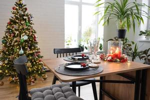 Festive table set in the living room for Christmas and New year in loft style. Christmas tree, black plates and forks, woven napkins, trendy tableware, cozy interior of the house photo