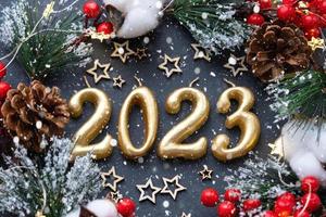 The golden figures 2023 made of candles on a black stone slate background are decorated with a festive decor of stars, sequins, fir branches, balls and garlands. Greeting card, happy New Year. photo