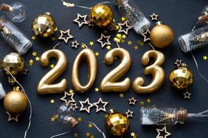 The golden figures 2023 made of candles on a black stone slate background are decorated with a festive decor of stars, sequins, fir branches, balls and garlands. Greeting card, happy New Year. photo