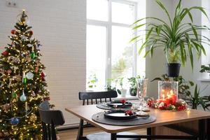 Festive table set in the living room for Christmas and New year in loft style. Christmas tree, black plates and forks, woven napkins, trendy tableware, cozy interior of the house