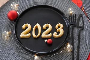 Golden figures 2023 from candles on a black plate on a festive table with a New Year's serving. Loft-style interior, party, feast. Wicker napkin, fork, knife photo