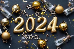 las figuras doradas 2024 hechas de velas sobre un fondo de pizarra de piedra negra están decoradas con una decoración festiva de estrellas, lentejuelas, ramas de abeto, bolas y guirnaldas. tarjeta de felicitación, feliz año nuevo. foto