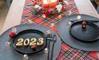 Golden figures 2023 from candles on a black plate on a festive table with a New Year's serving. Loft-style interior, party, feast. Wicker napkin, fork, knife photo