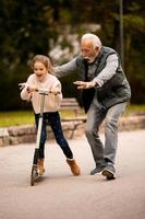 Senior man teaching his granddaughter how to ride kick scooter in park photo