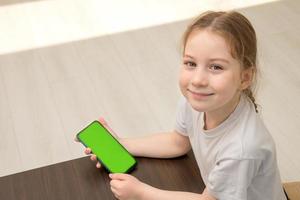 little girl sits at a table holding a phone with a green screen in her hands, top view photo