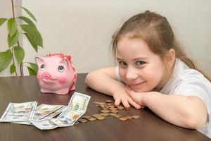 there is a piggy bank on the table, money lies nearby, a smiling baby sits at the table photo